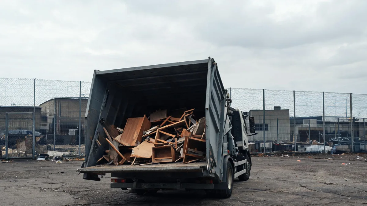 Ciężarówka z otwartą skrzynią pełną połamanych płyt i drewnianych mebli, na betonowym placu za siatką