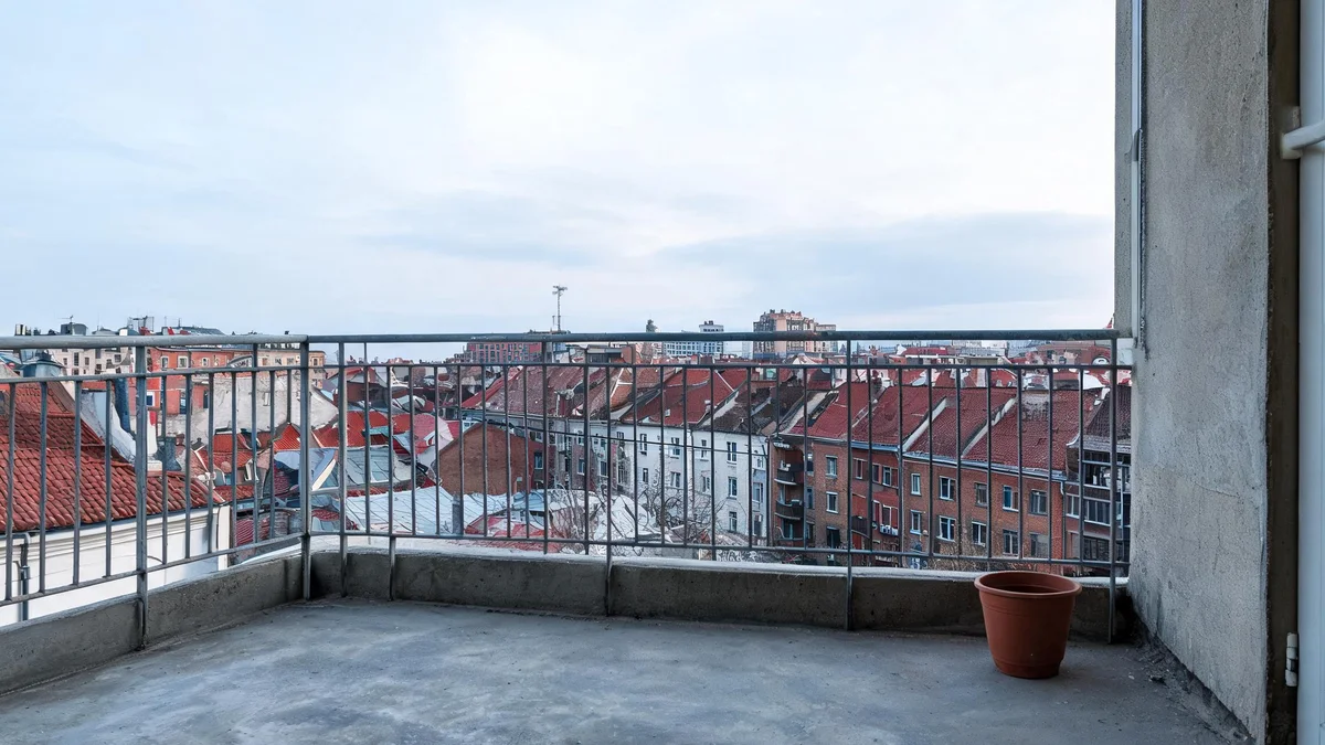 Pusty balkon z betonową posadzką i stalową balustradą, w rogu jedna donica, w tle dachy kamienic